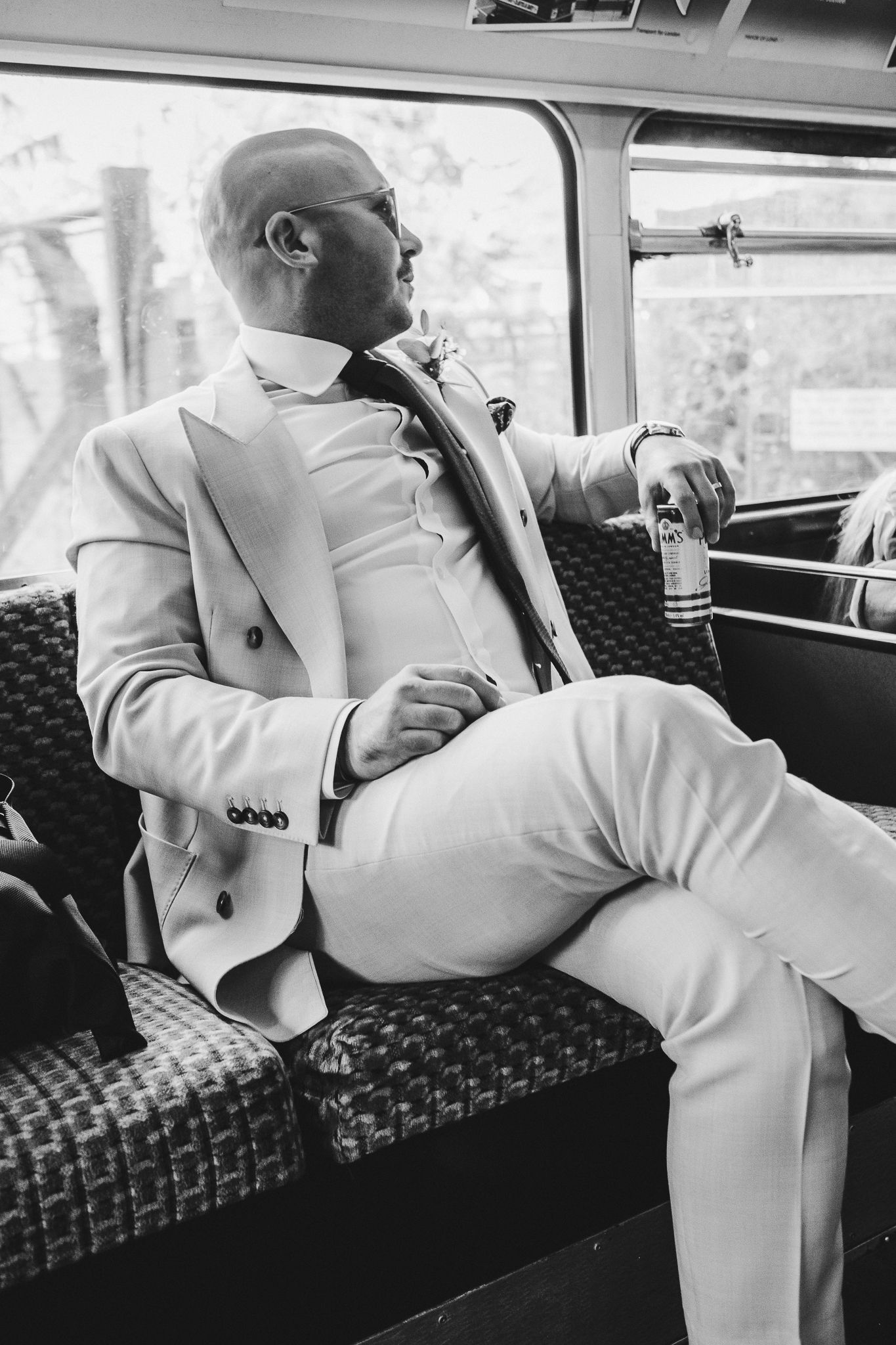 A person in a white suit and sunglasses relaxes on patterned vintage bus seating with a drink in hand.