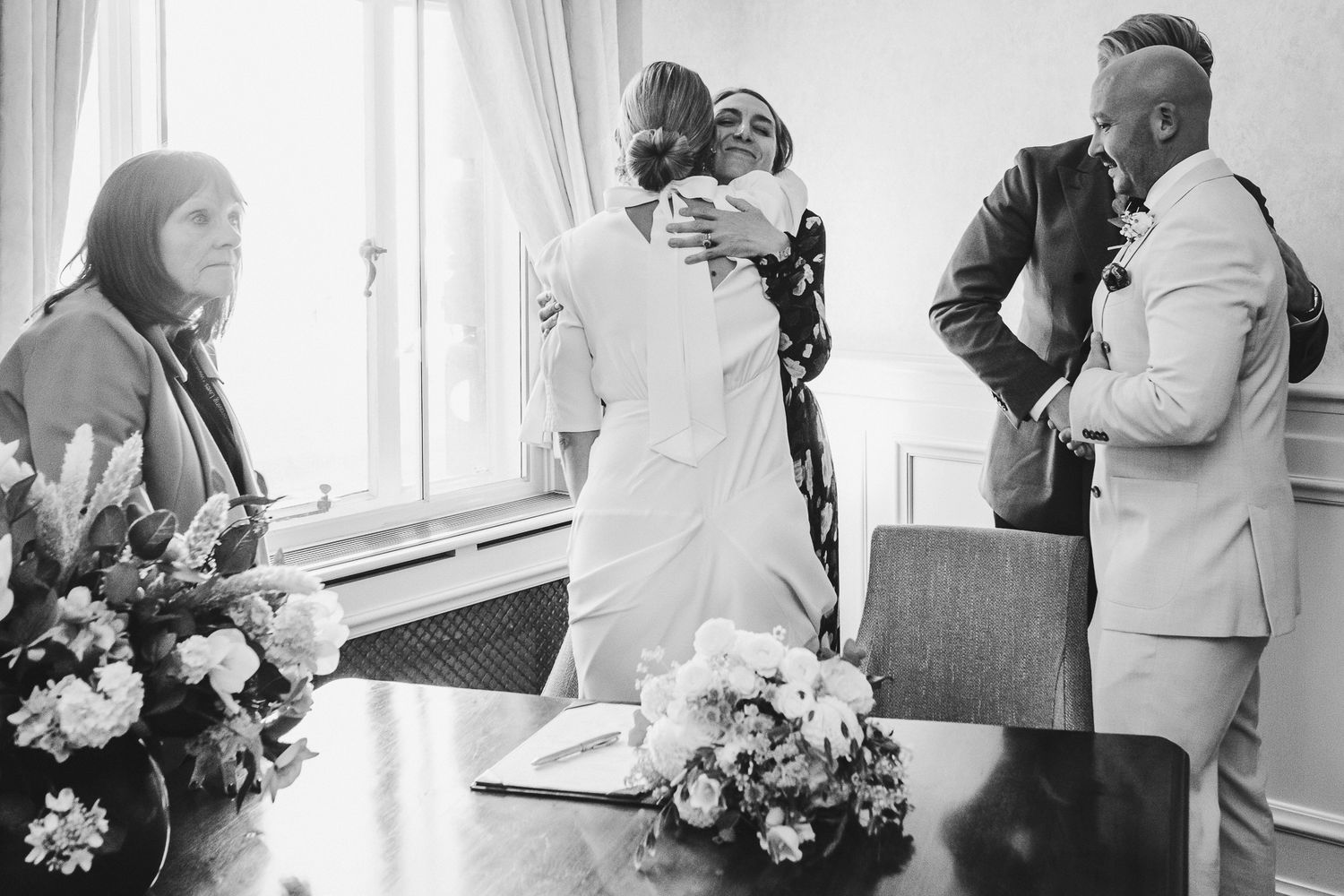 Black and white photograph showing a wedding ceremony gathering with floral arrangements in a bright interior setting.