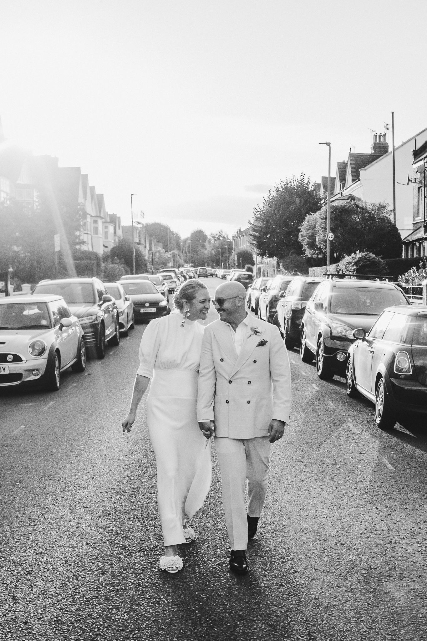 Two individuals in white paint-splattered coveralls walk together on a paved road lined with parked cars on a bright day.