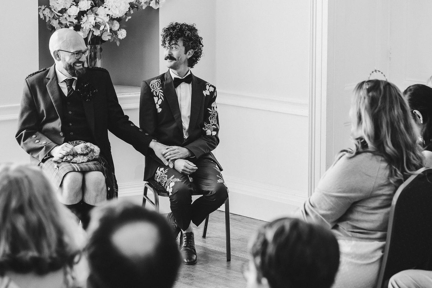 Two grooms in dark suits hold hands while seated during an intimate wedding ceremony as guests look on in a bright, elegant room.