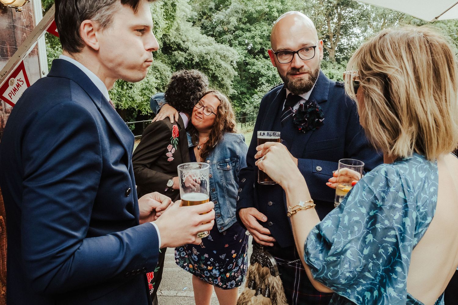 Wedding guests in formal attire chat and hold drinks together outdoors, with a bearded man in a navy suit and boutonniere smiling nearby.