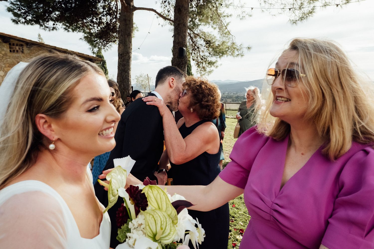 Smiling bride in a white dress holds a bouquet of white calla lilies while greeting a woman in a pink top at an outdoor wedding.