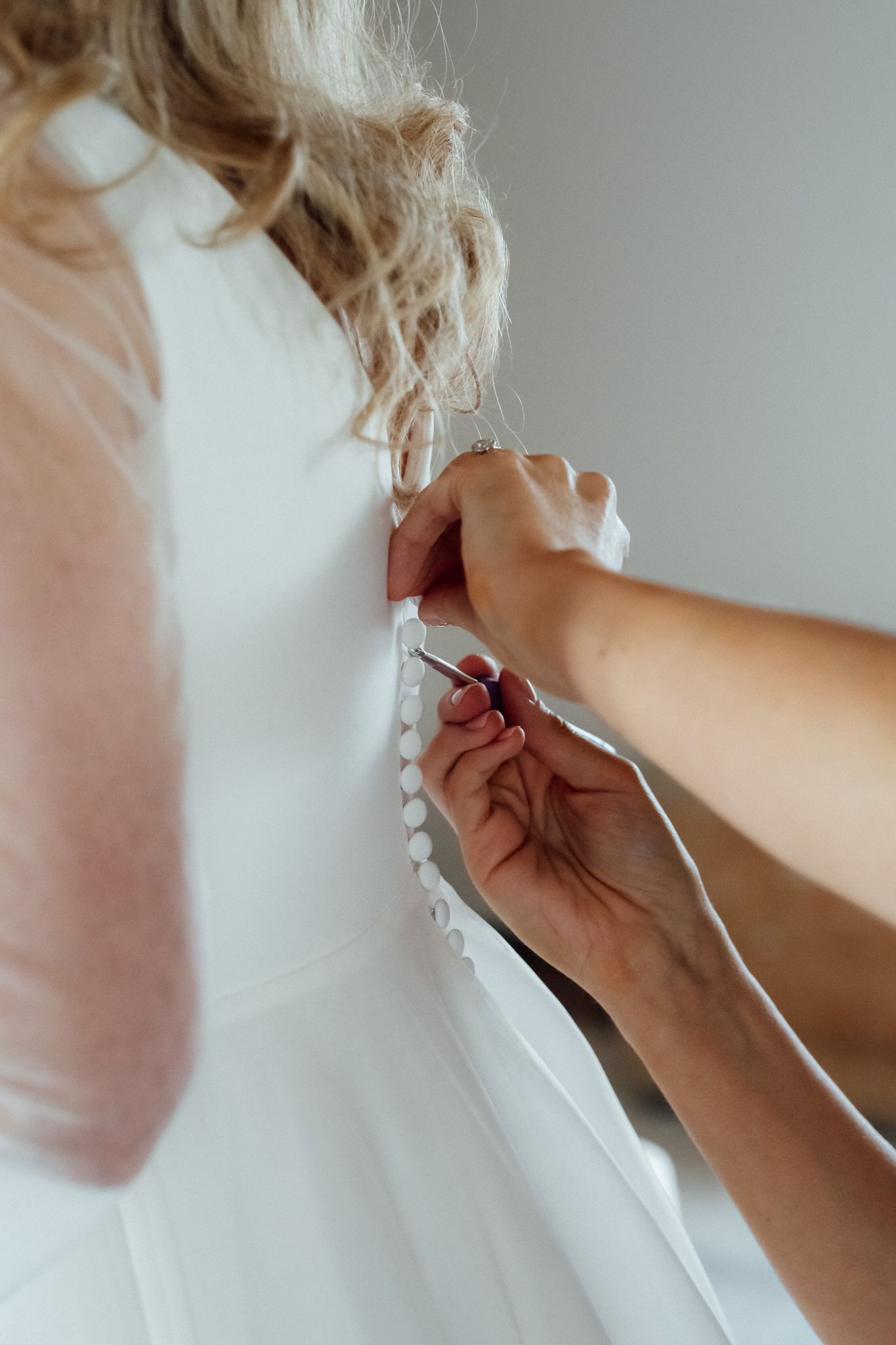 Bridesmaid fastening the small covered buttons on the back of a white wedding dress on the bride, with loose blonde curls.