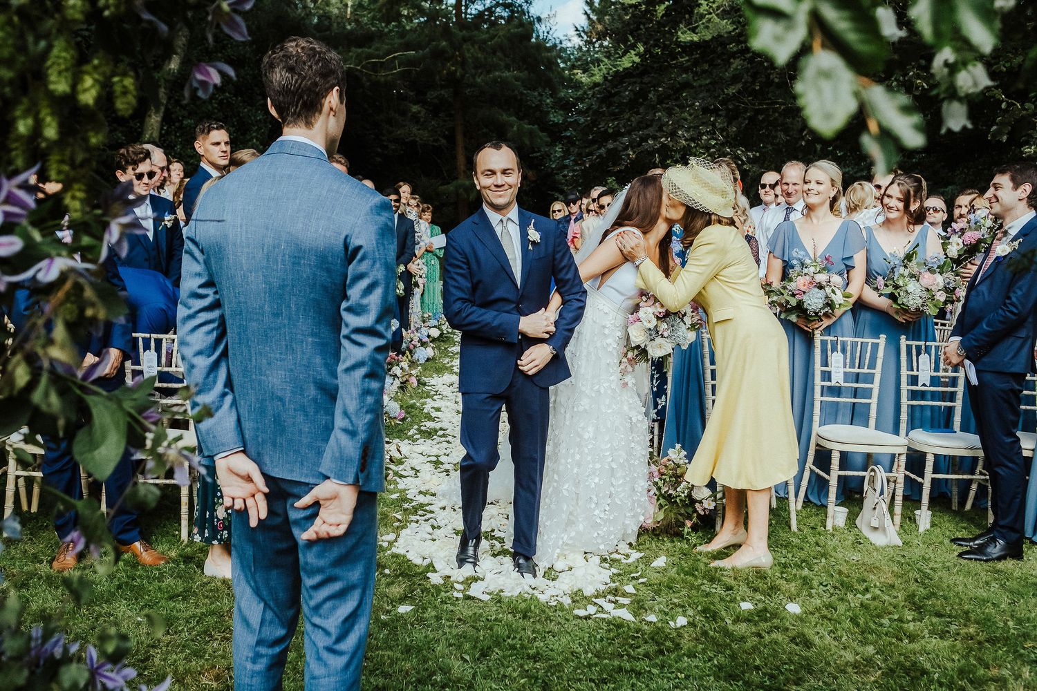 Outdoor wedding ceremony setup with white chairs and guests gathered among trees.