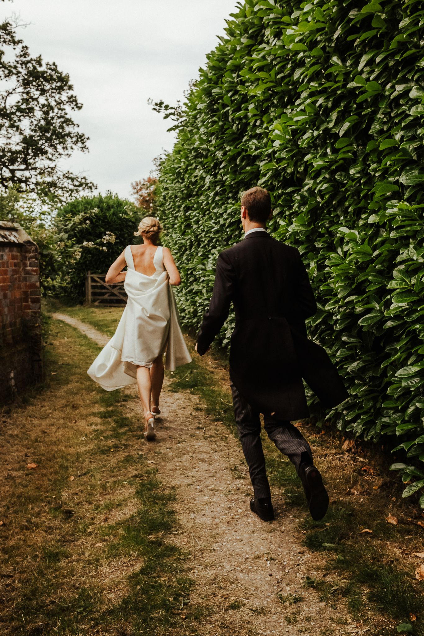 A couple walking down a garden path lined with tall green hedges on their wedding day.