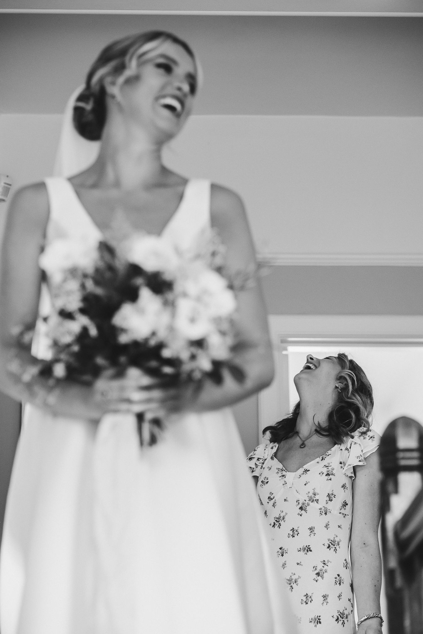 Black and white photo of a bride in a white dress holding a bouquet with a flower girl laughing.
