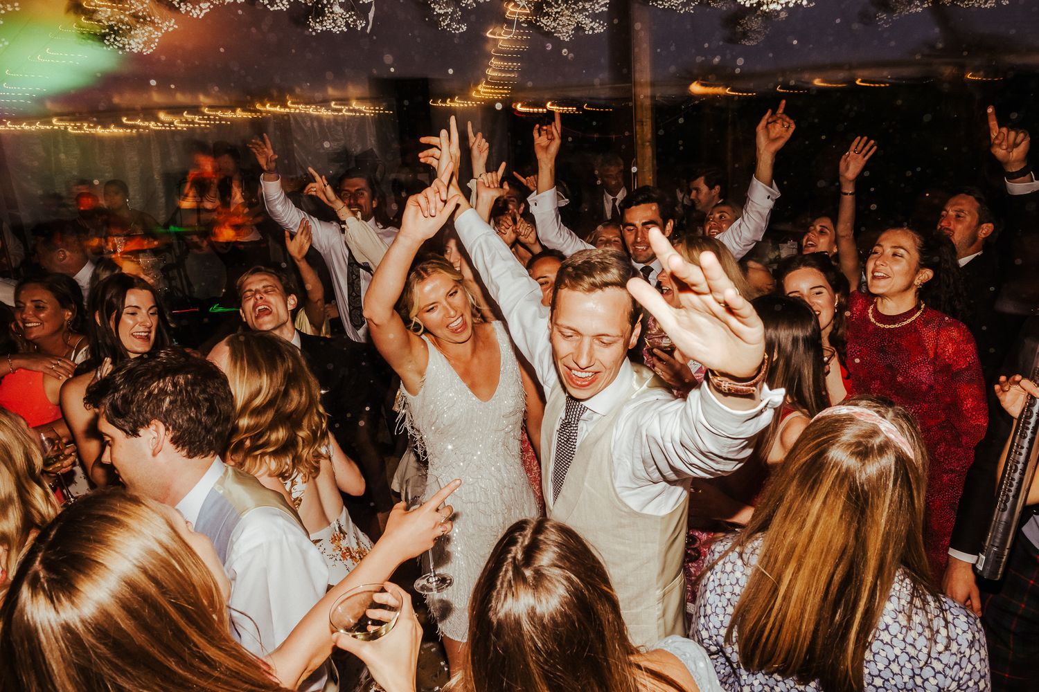 Energetic crowd dancing and celebrating at a wedding reception under string lights.