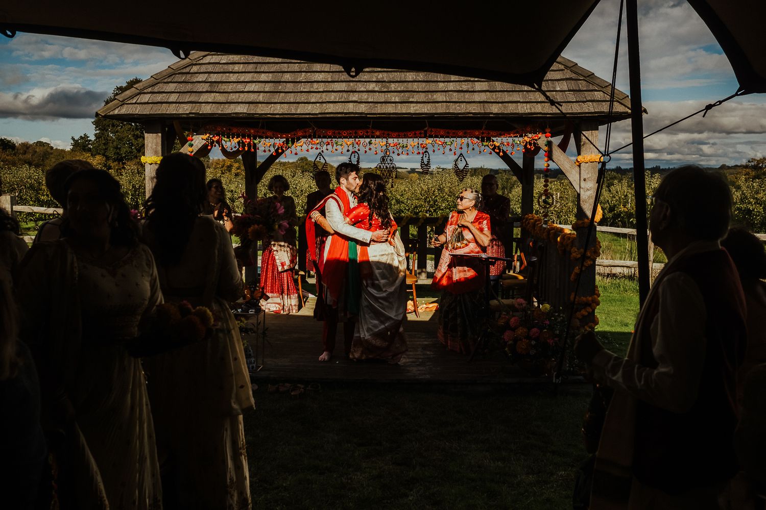 Silhouettes of wedding guests gather under a wooden gazebo during sunset.
