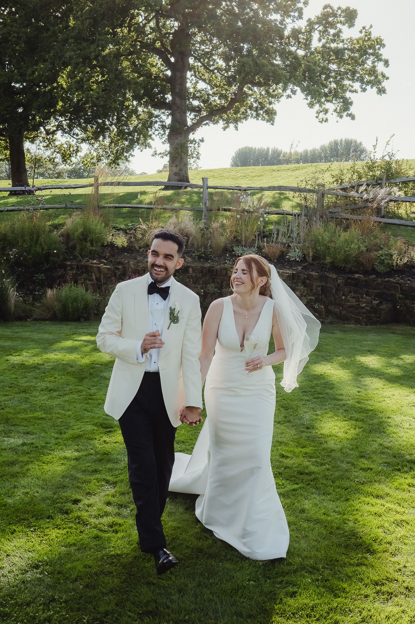 A couple in wedding attire shares joyful moments together on a sunny lawn with trees in the background.