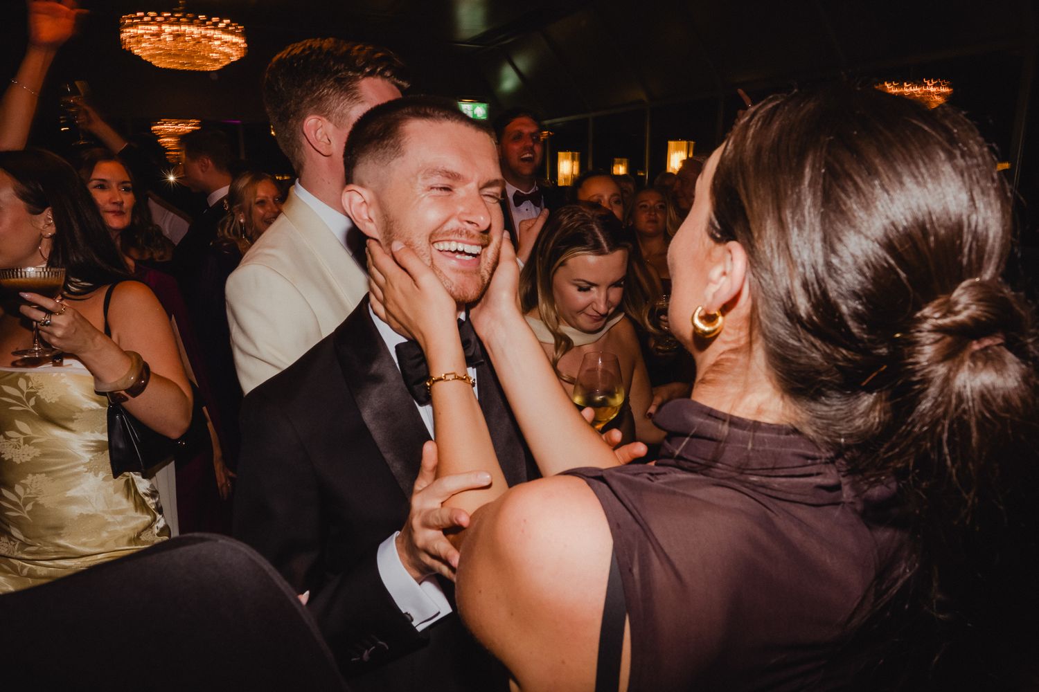 Wedding guests dancing and celebrating together on a dimly lit dance floor with warm lighting overhead.