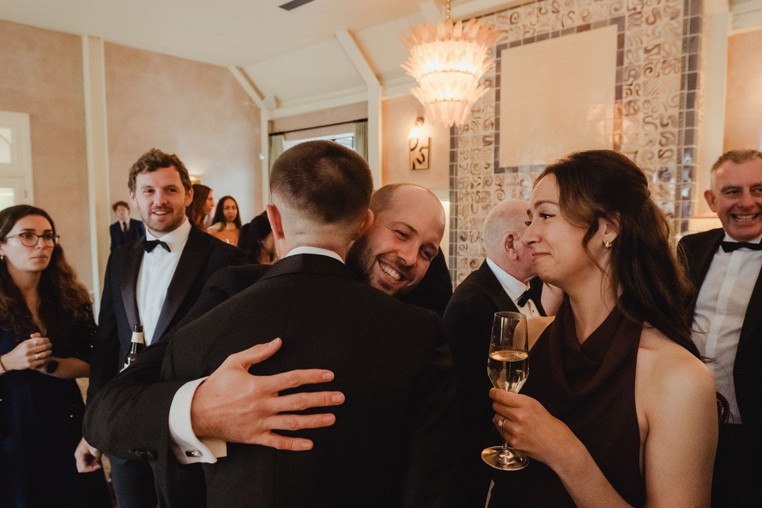 Guests mingle and embrace at an elegant indoor reception with exposed brick walls and warm lighting.