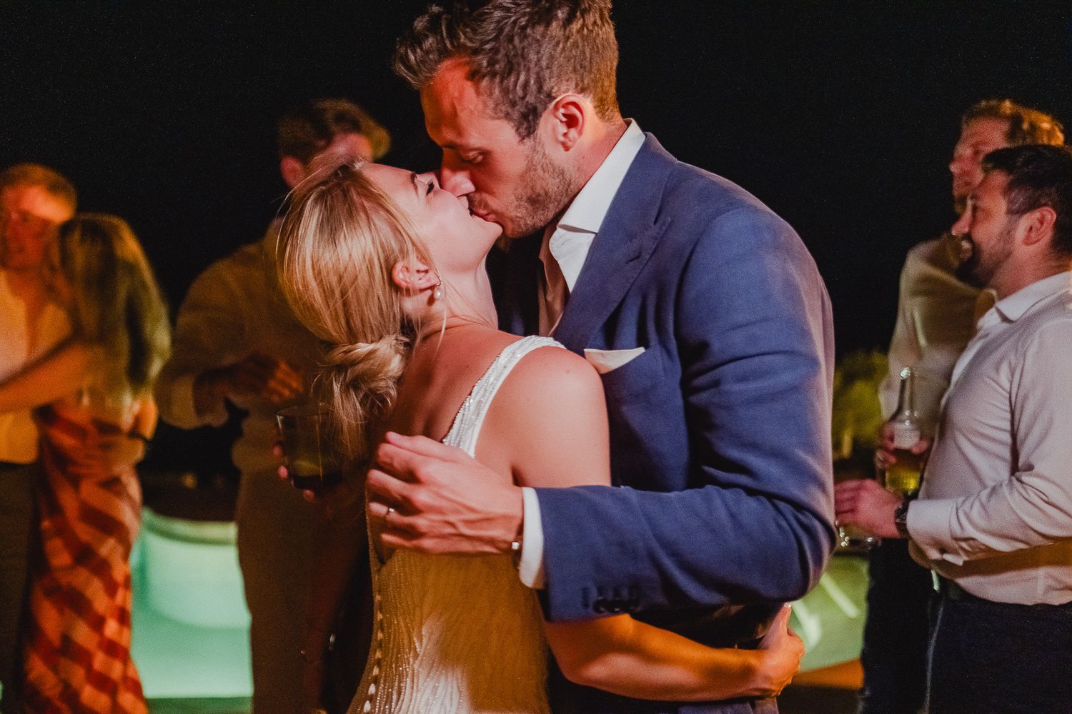 A couple shares a romantic dance moment at a wedding reception with guests and warm lighting in the background.