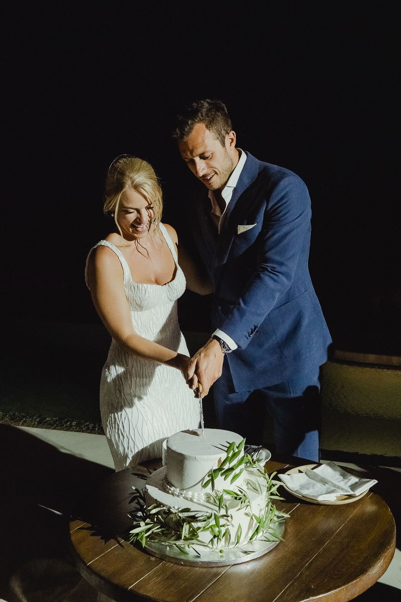 A couple in wedding attire cuts their wedding cake together in dramatic lighting at their reception.