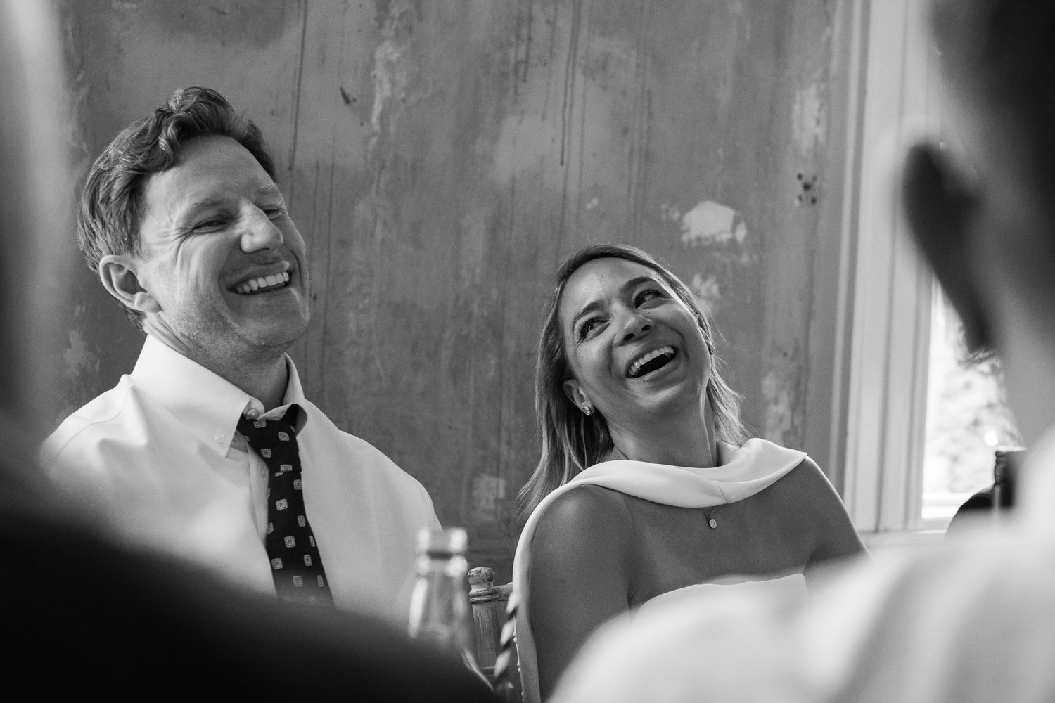Black and white photo of a smiling man in a tie and a laughing woman with her head tilted back, enjoying a joyful moment at an event.