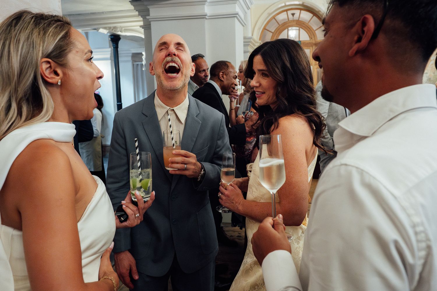 Guests laugh and toast with champagne glasses at a lively indoor celebration, with a bald man in a gray suit laughing in the center.