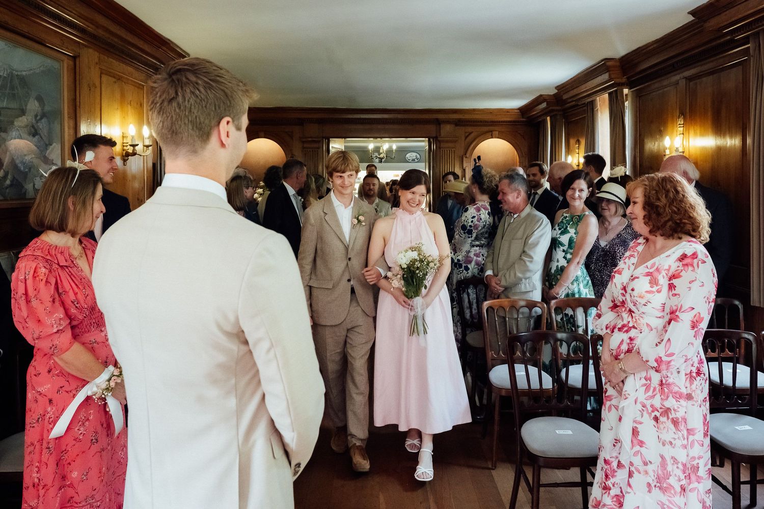 Bride in a pink dress carrying a bouquet walks down the aisle toward the groom as wedding guests watch in a wood-paneled room.