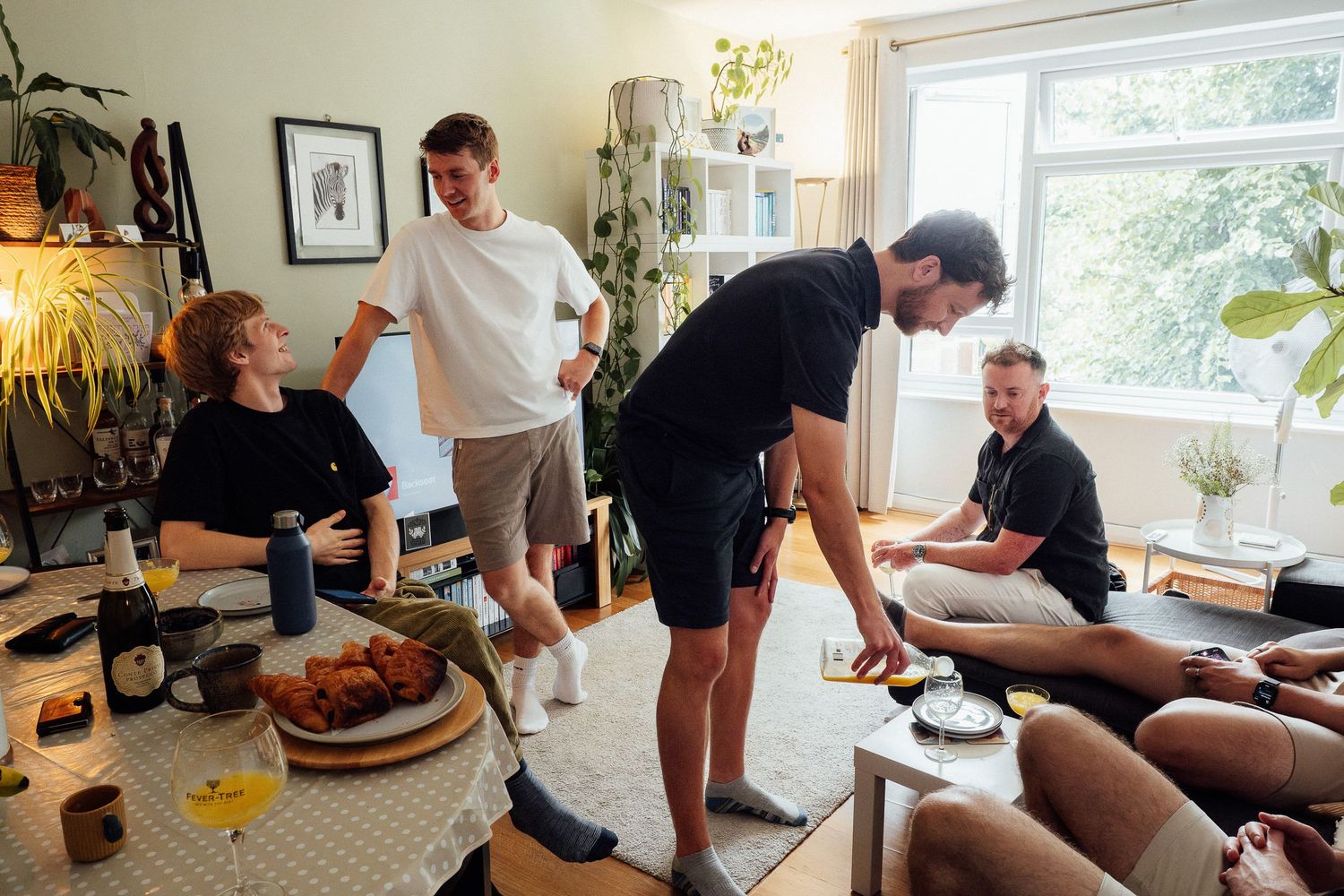 Friends gather and chat in a bright living room around a table with brunch food, drinks and houseplants on a sunny day.