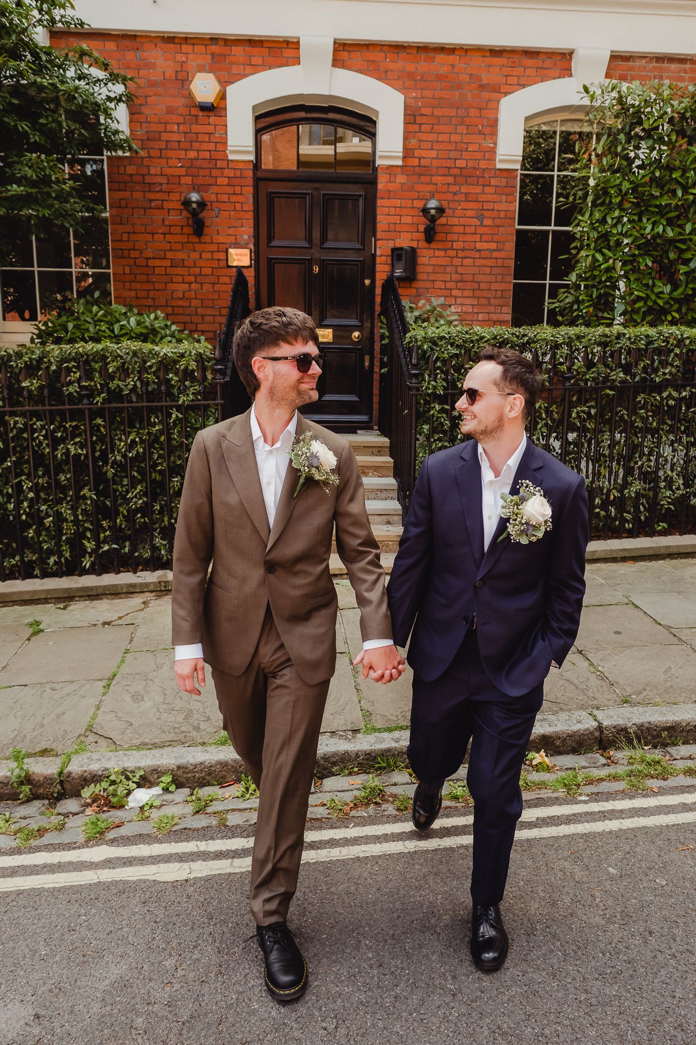 Two people in suits holding hands while walking on a sidewalk in front of a red brick building with white trim.