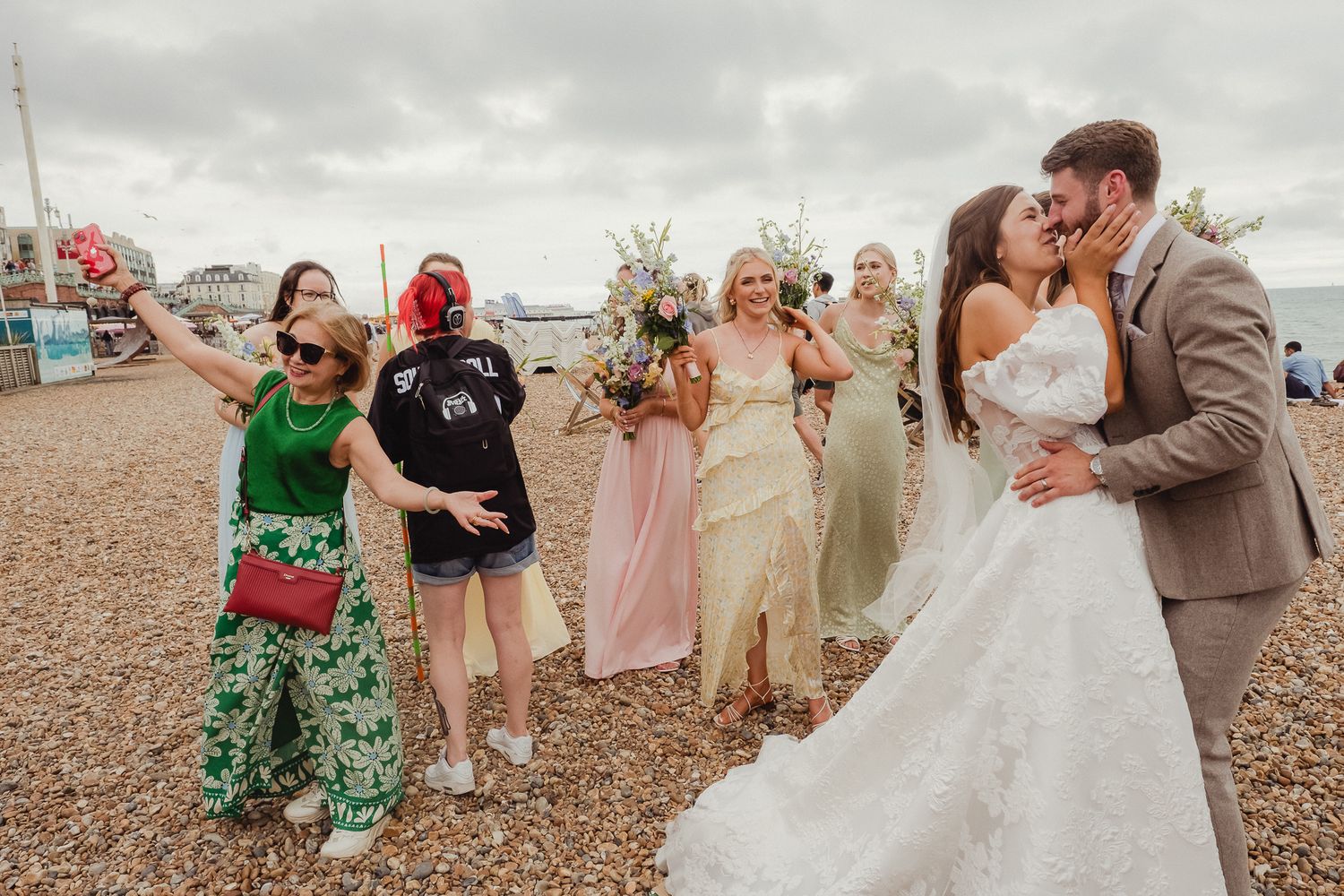 Wedding party celebrates joyfully on a pebble beach with bridesmaids in pastel dresses and groomsmen in suits.