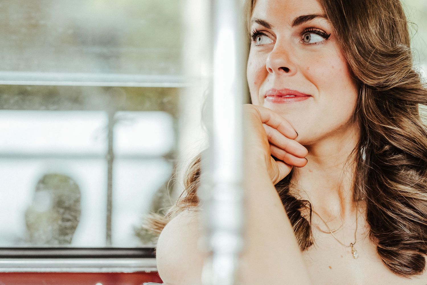 Woman with wavy brown hair and natural makeup gazes thoughtfully to the side near a window in soft lighting.