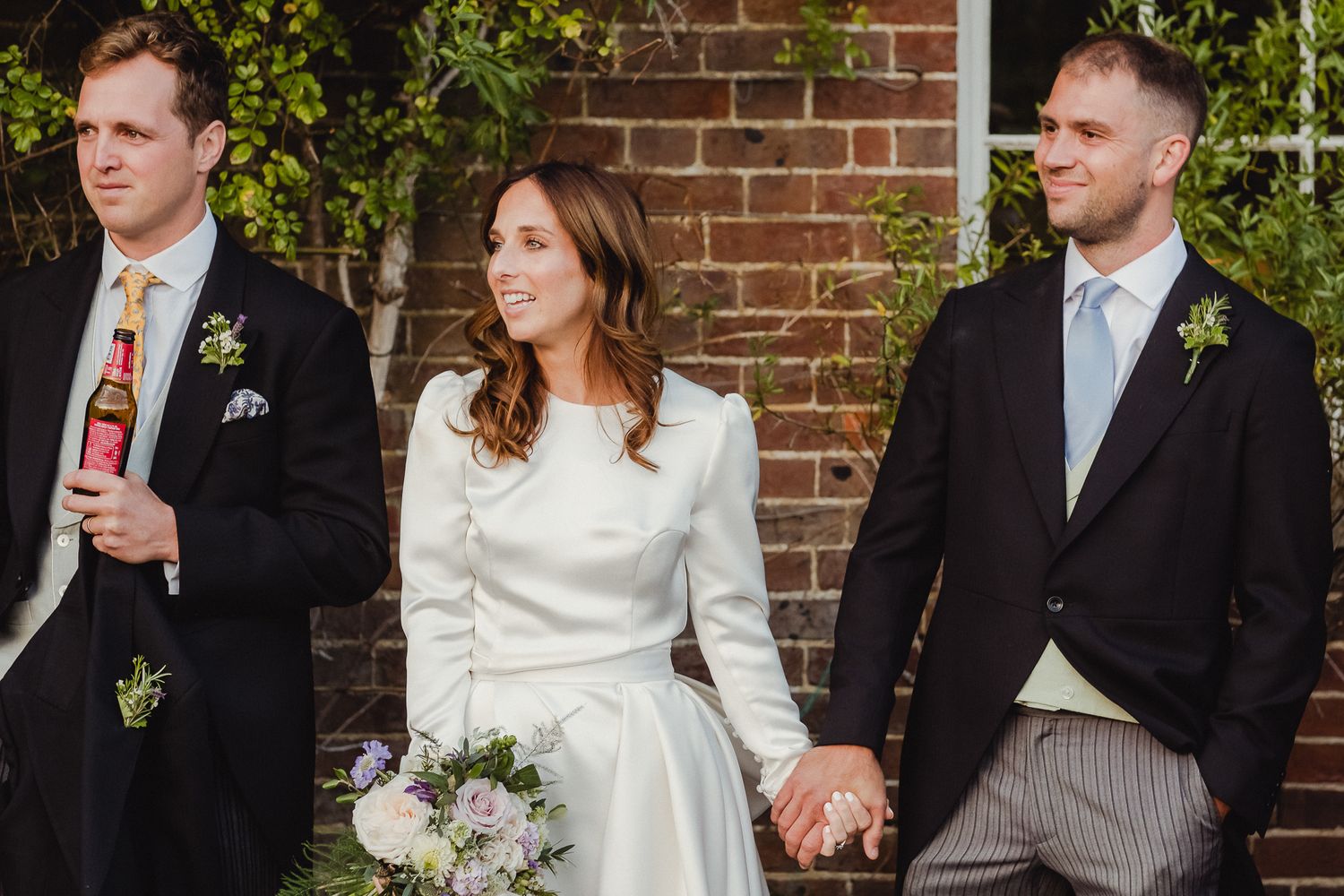 A wedding party in formal attire stands together against a brick wall backdrop with greenery.