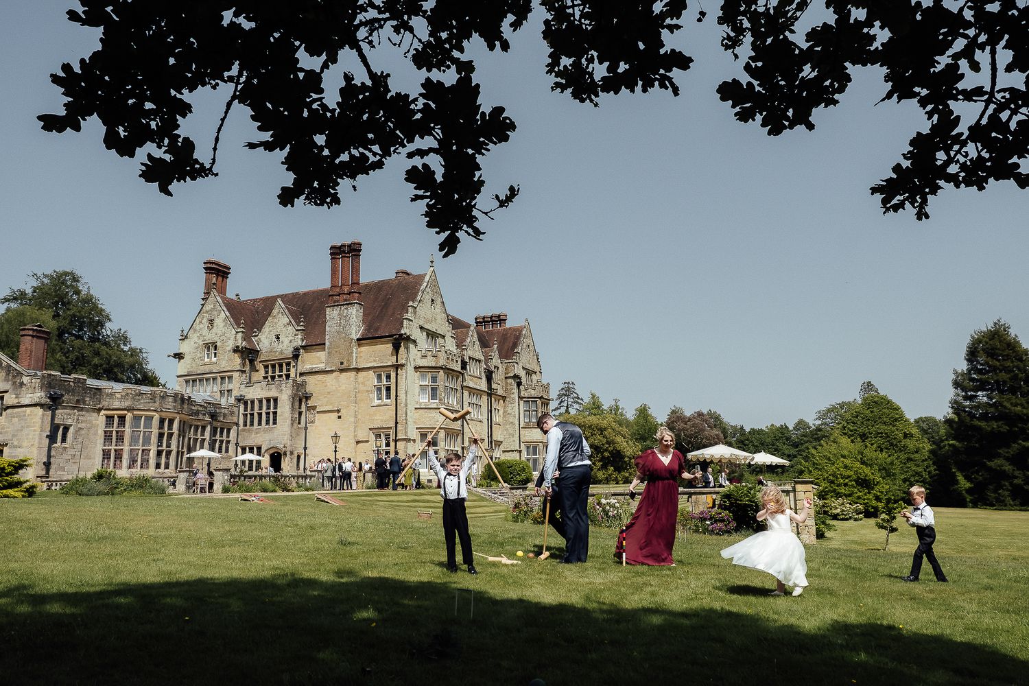 Wedding party celebrating on lush green lawn outside a grand historic English manor on a sunny day.