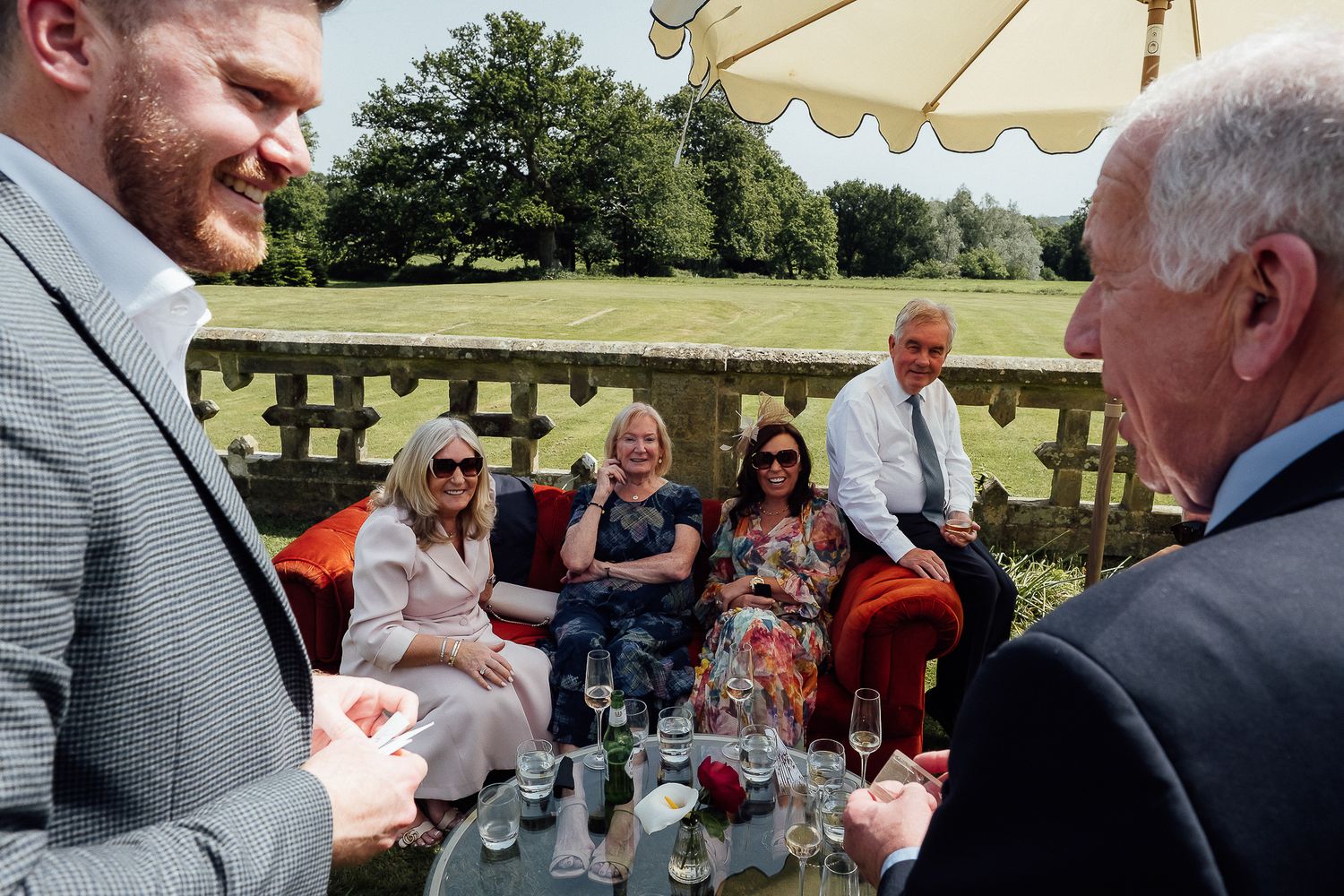 Guests relaxing on red sofas at an outdoor garden party, enjoying drinks under a striped umbrella.