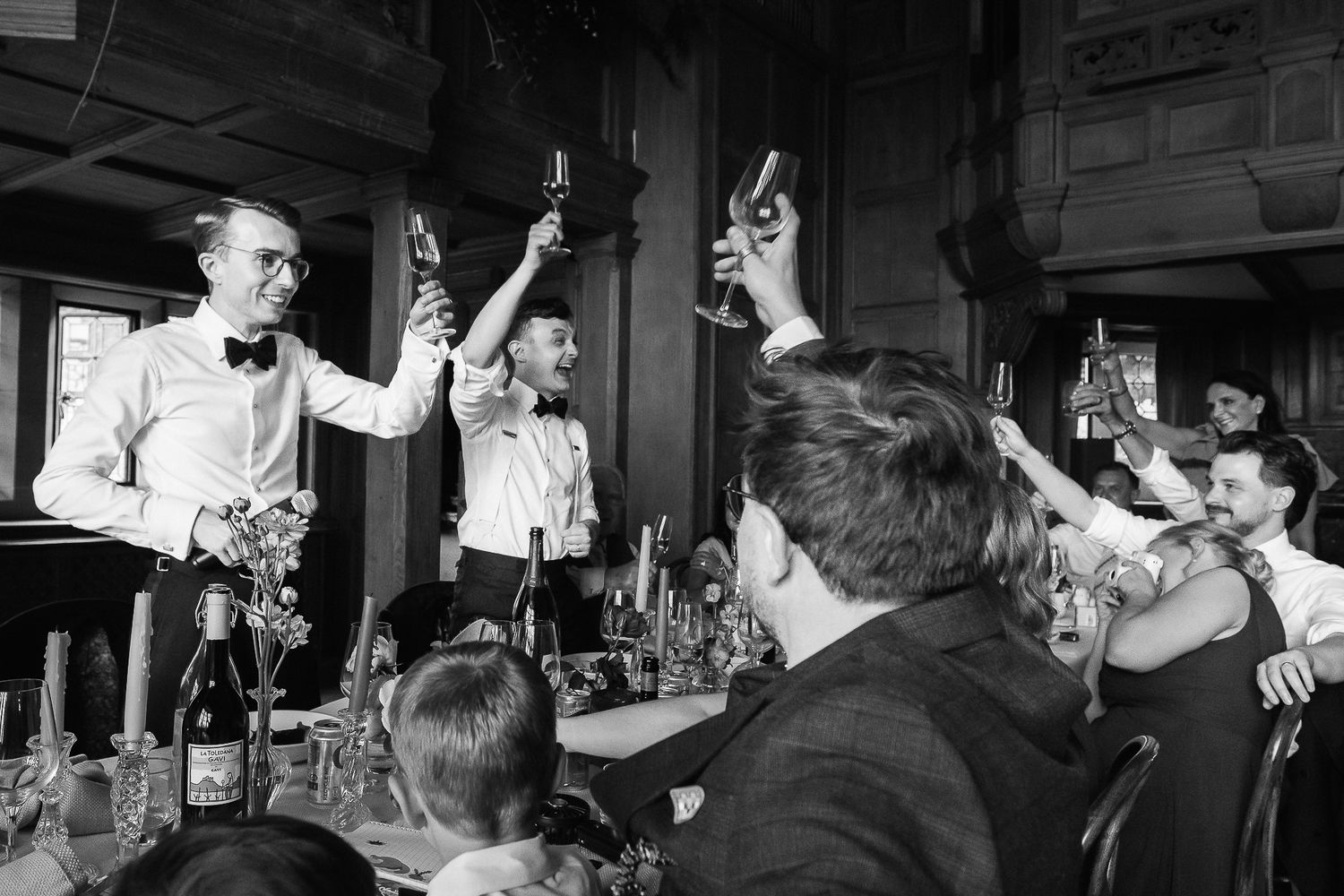 Black and white photo of elegantly dressed guests raising champagne glasses in a toast at a formal wedding reception.