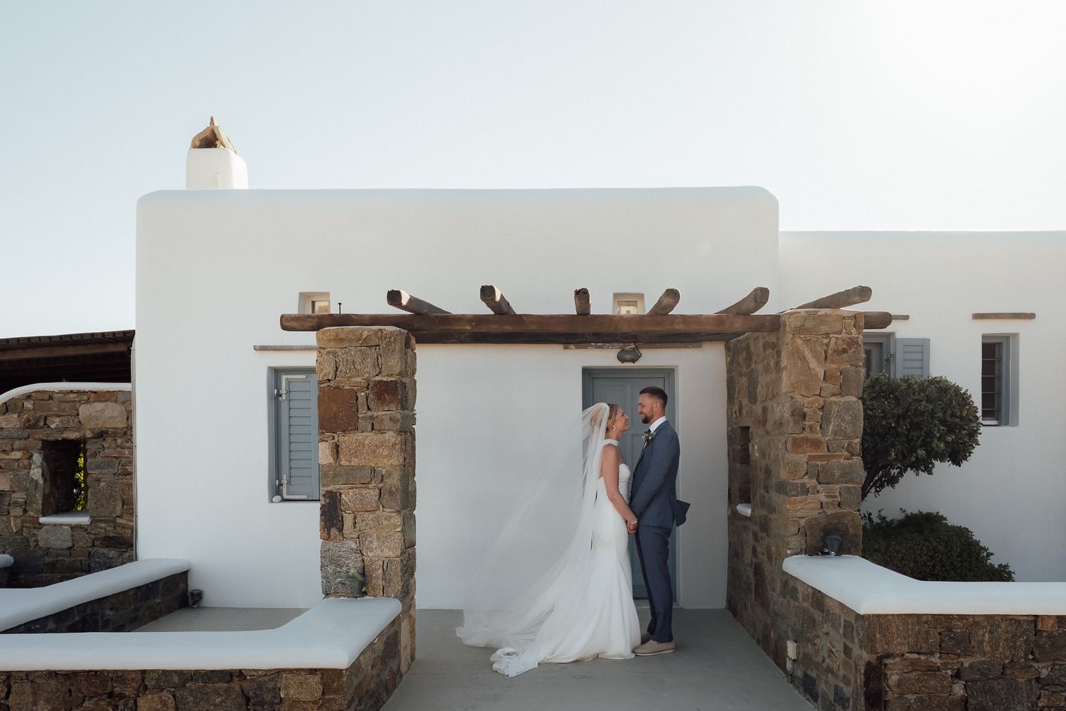 Bride and groom share a romantic moment under a rustic wooden arch at a whitewashed Mykonos wedding venue.