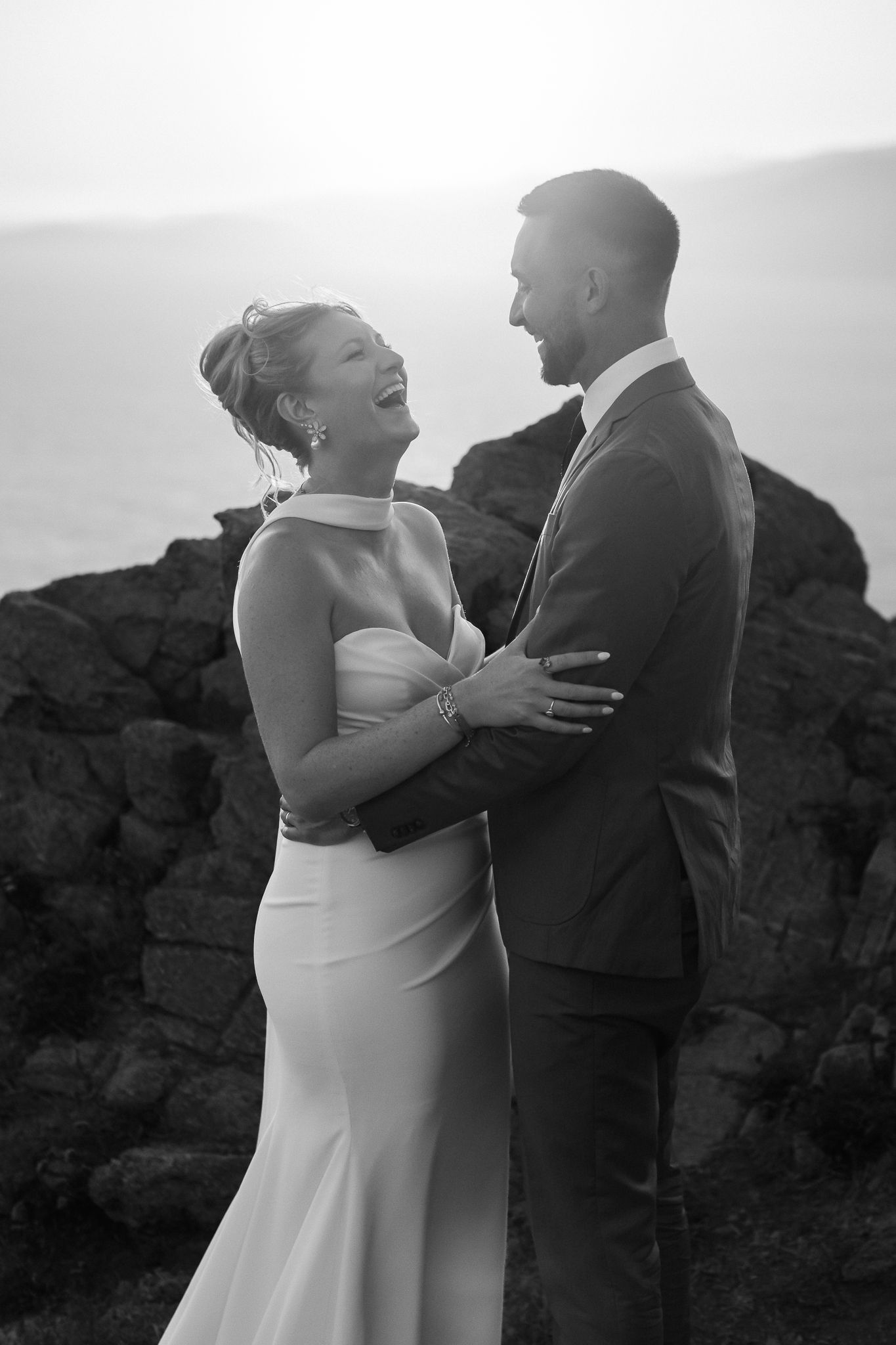 Black and white photo of a laughing bride in a white gown embracing a suited groom outdoors at sunset.