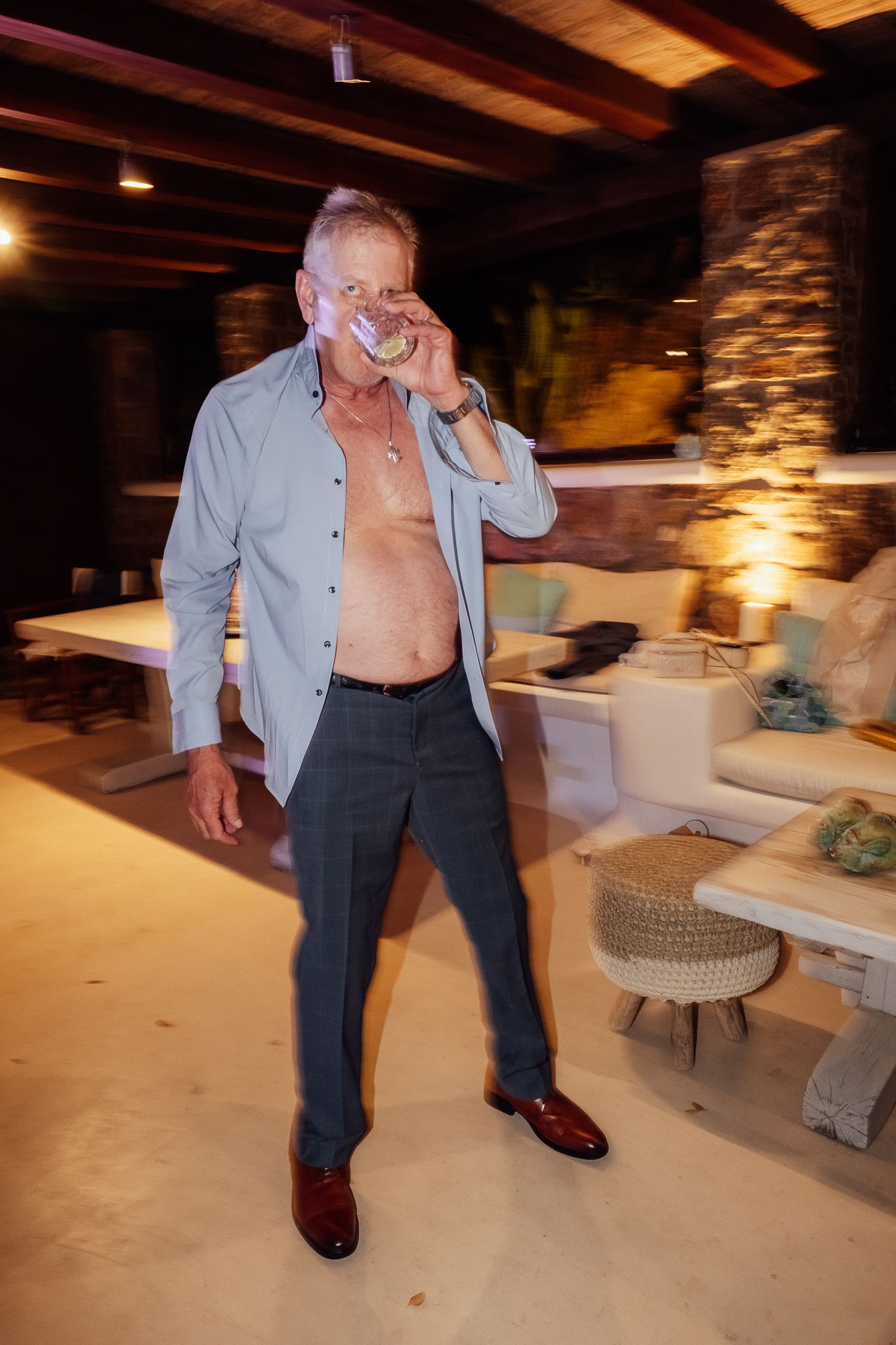 Older man with open shirt drinking from glass in dimly lit luxury lounge with stone walls.