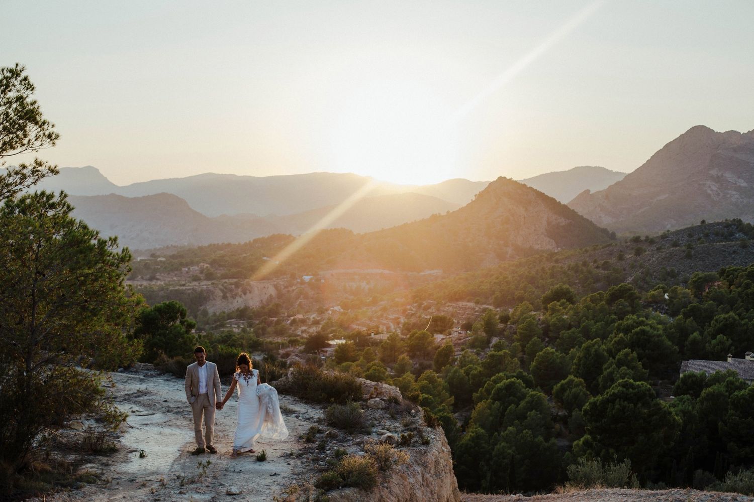 A couple stands on a rocky overlook during sunset with mountains in the distance.