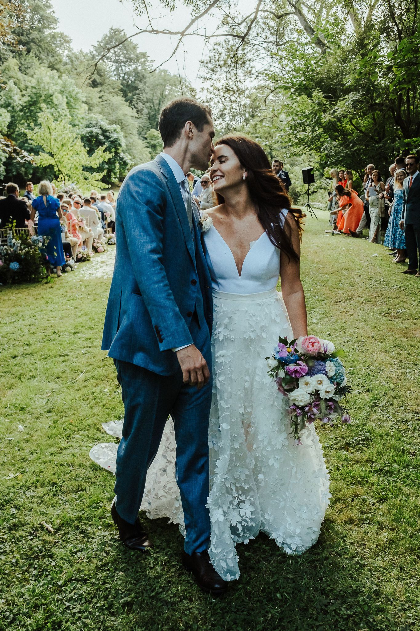 Newlyweds share intimate moment on garden lawn surrounded by wedding guests.