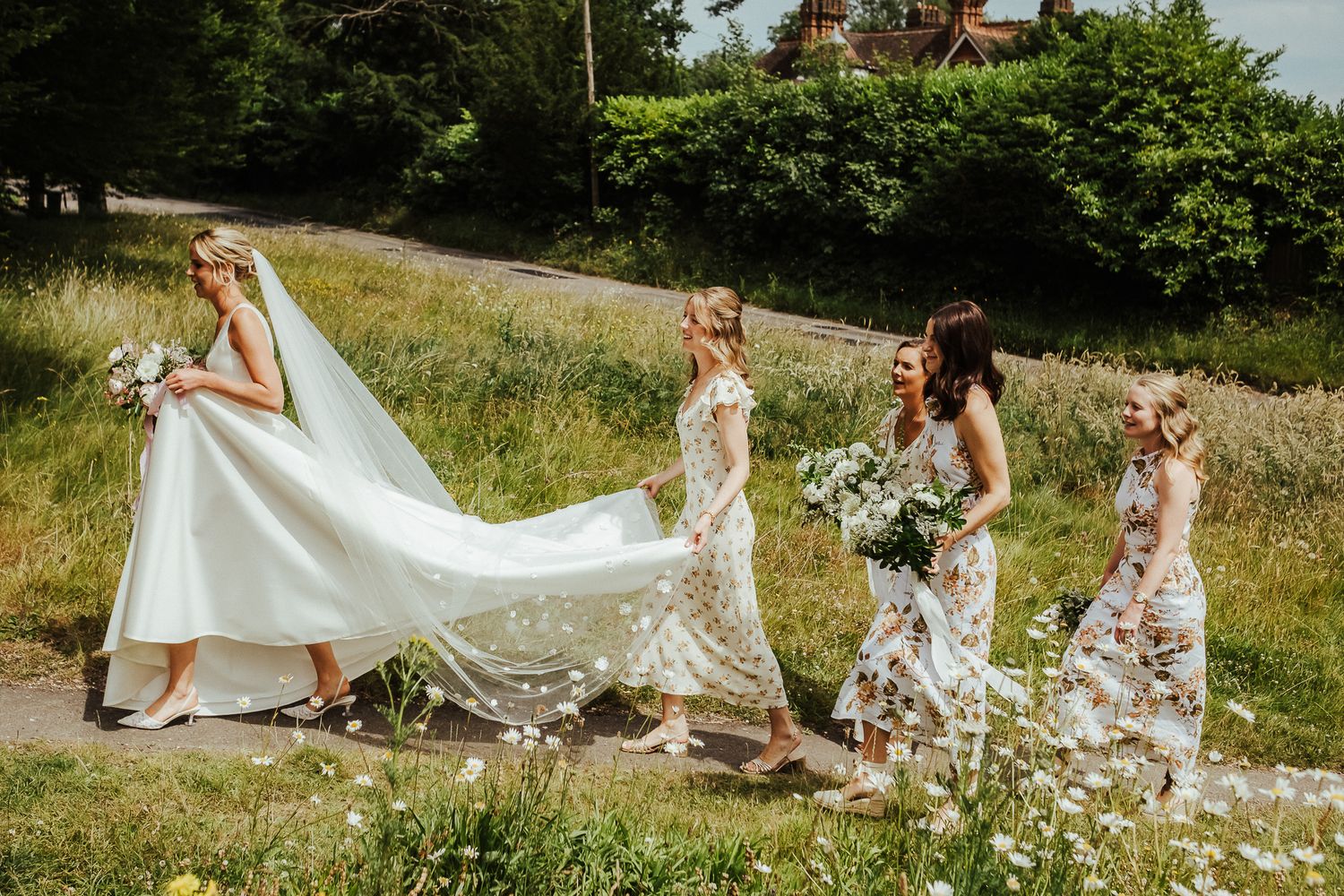 Wedding party members walk through a grassy field with flowing dresses and a trailing white veil in the sunlight.