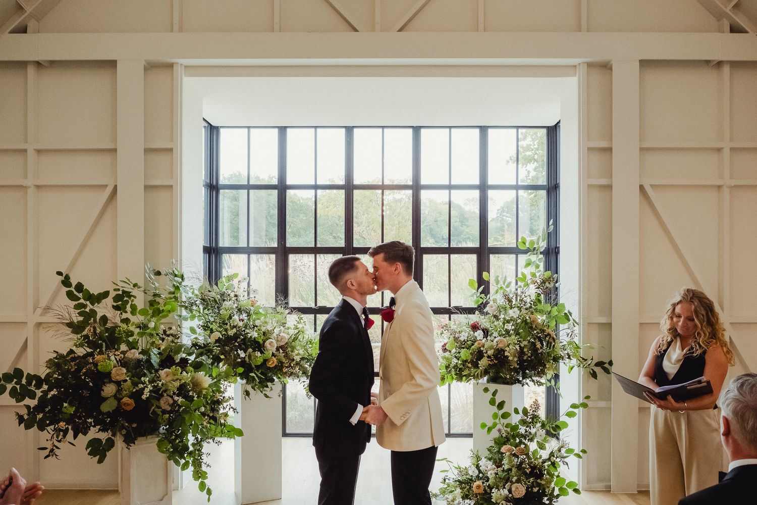 Two people share an intimate moment at the altar during a wedding ceremony with floral arrangements by a large window.
