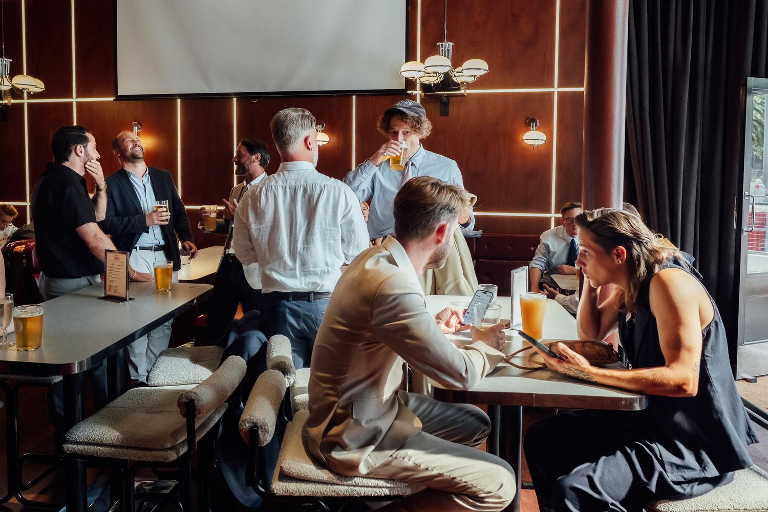 Group of people socializing over beer and drinks at tables in a warmly lit restaurant bar with wood-paneled walls and pendant lighting.