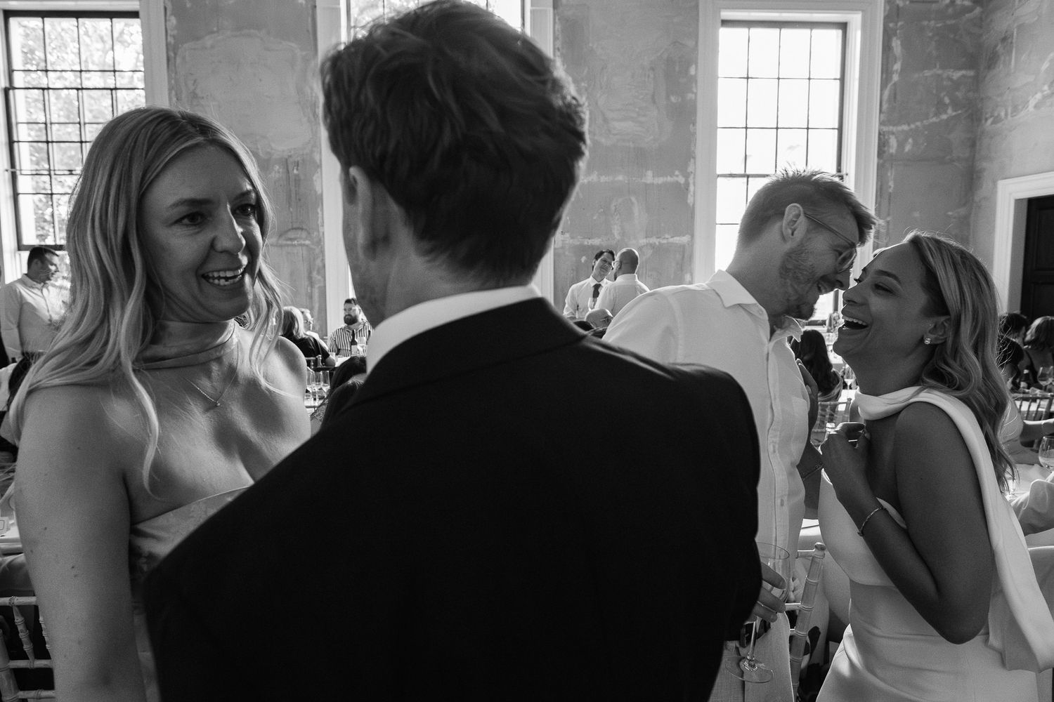Black and white photo of wedding guests laughing and chatting in a bright hall, with a man in a dark suit seen from behind.