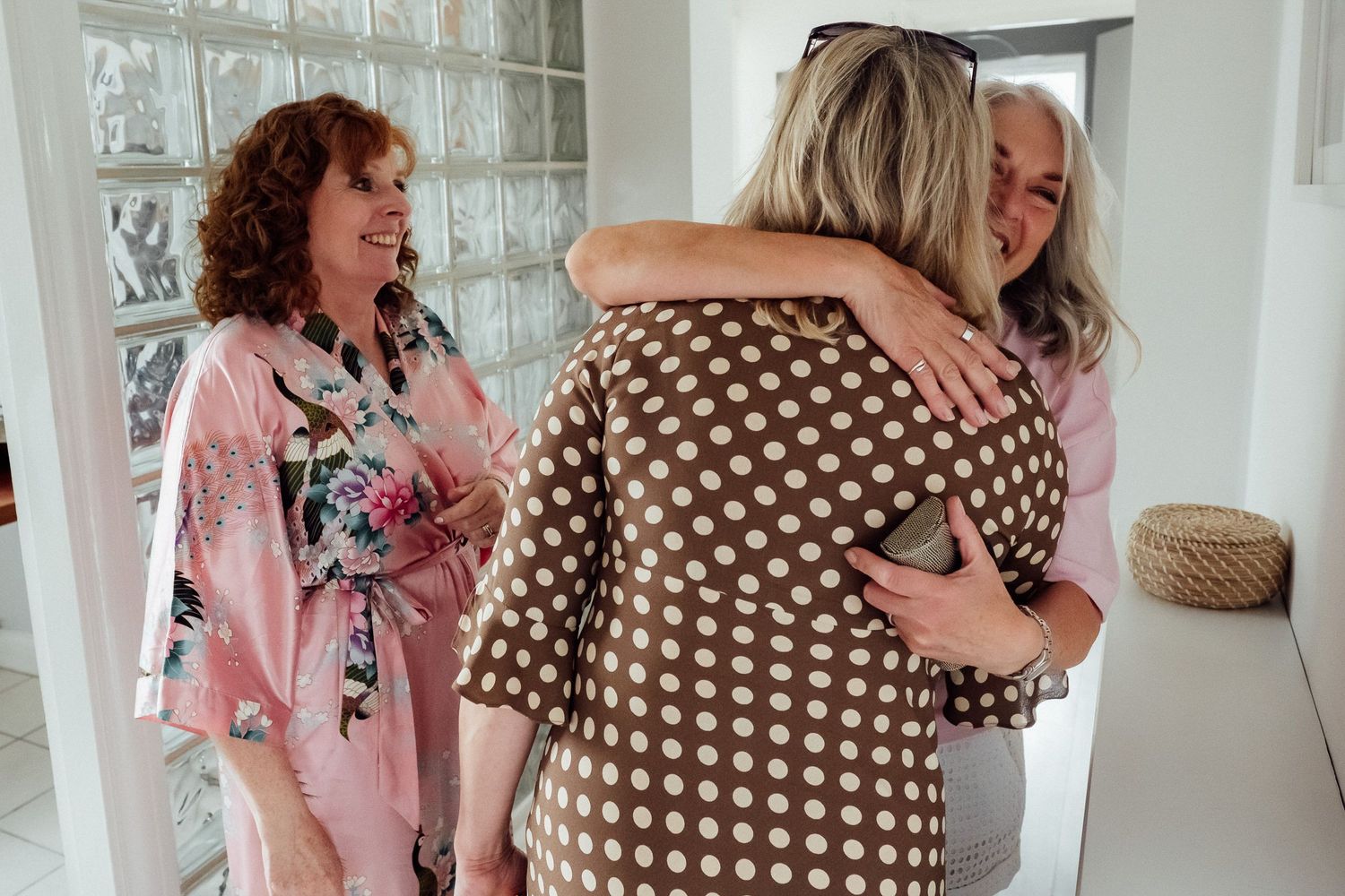 Two women share a warm hug in a bright hallway while a smiling woman in a pink floral robe looks on beside a glass block wall.