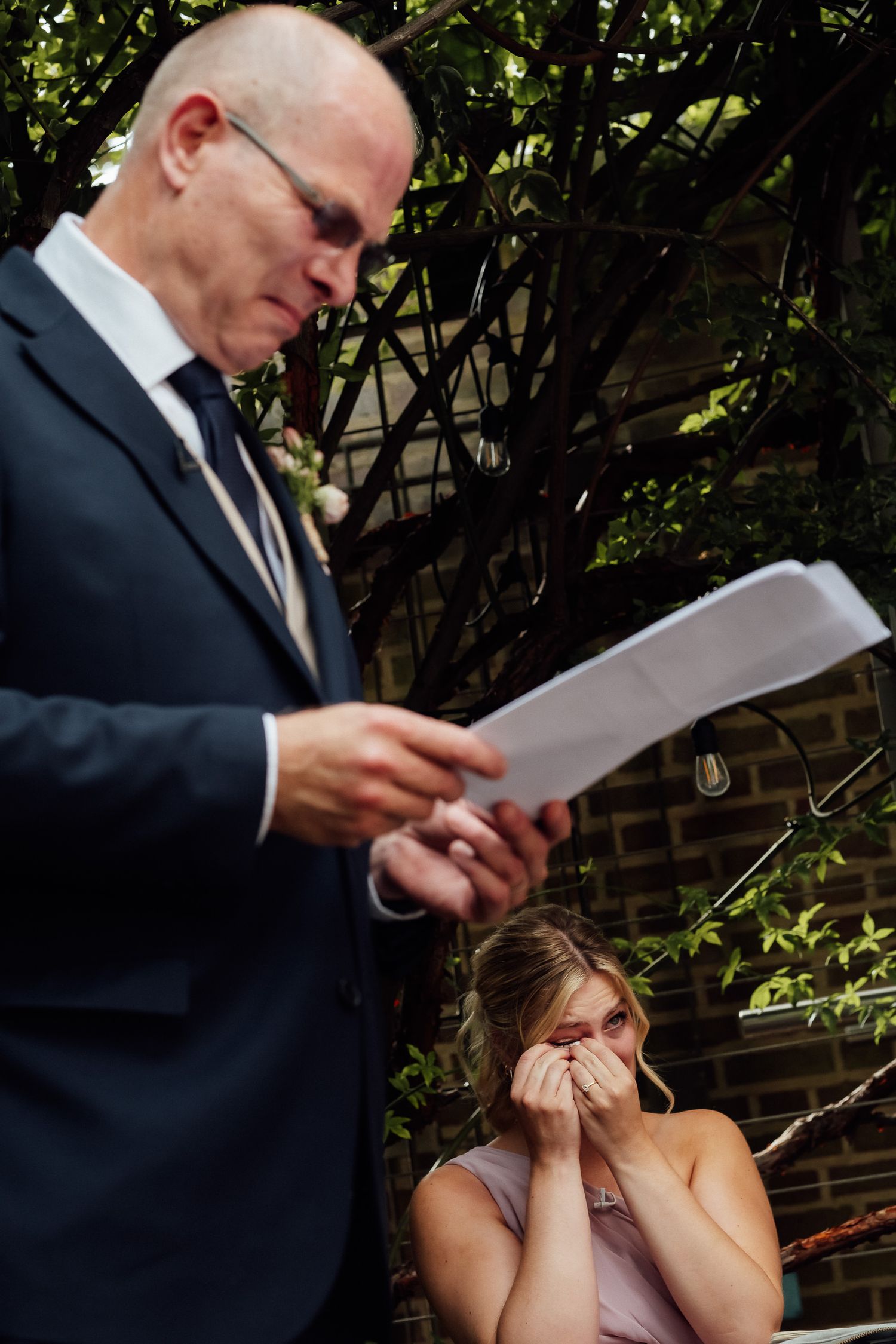 Emotional man in a navy suit reads from a paper during an outdoor ceremony while a woman in a blush dress wipes tears from her eyes.