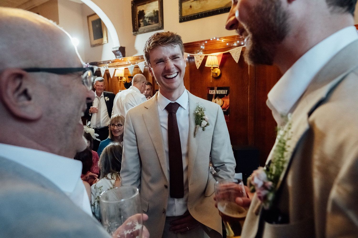 Smiling groom in a light suit with floral boutonniere laughs with guests during a lively wedding reception in a cozy pub.