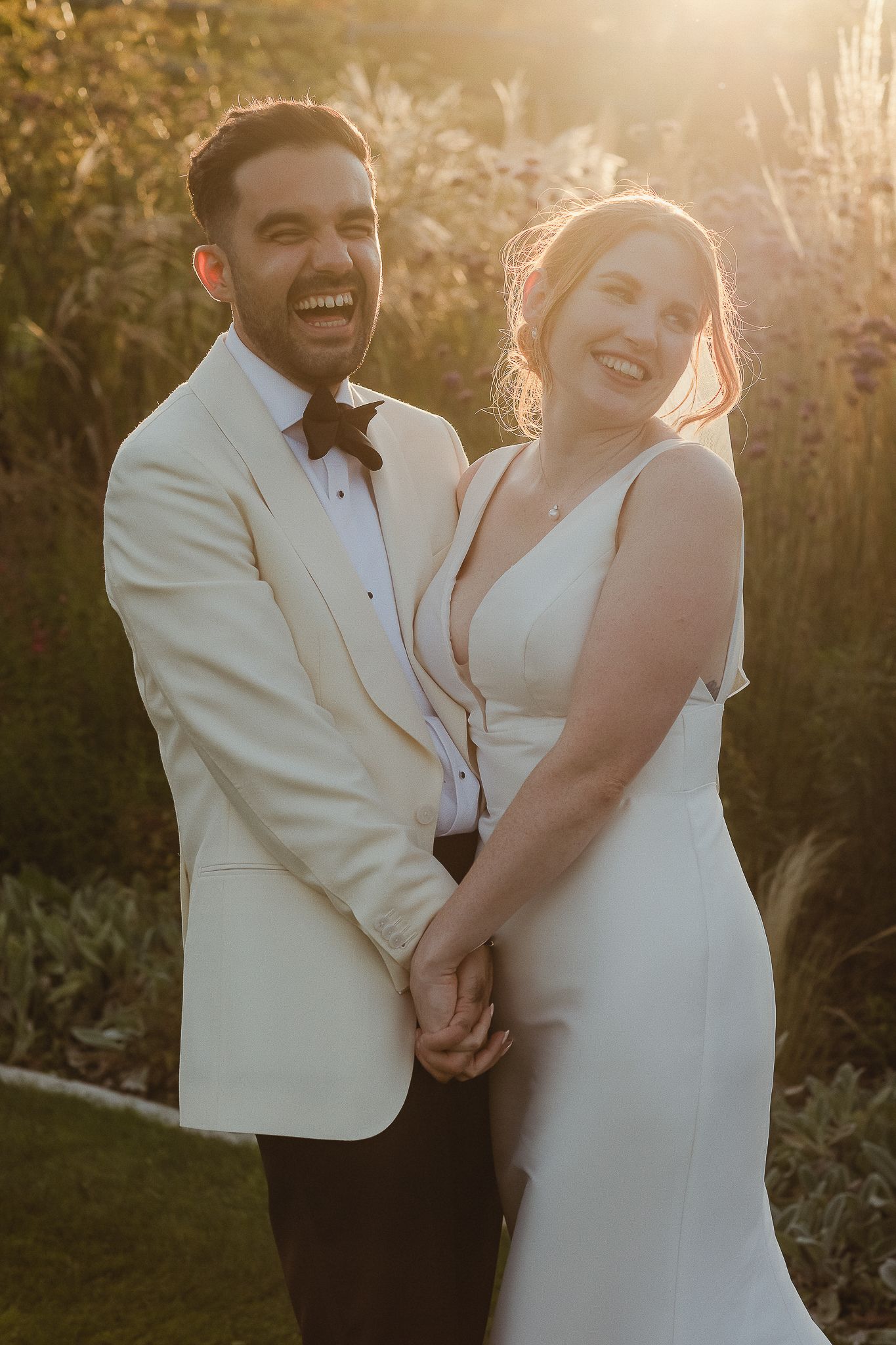 A joyful wedding couple laughs together in golden sunlight while wearing elegant white formal attire.