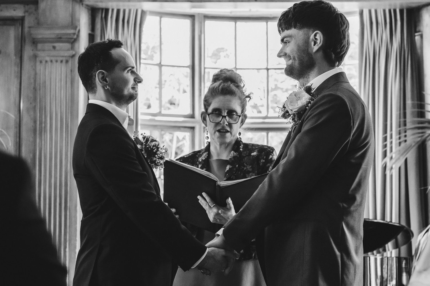 A black and white photograph of a wedding ceremony with two people holding hands in front of an officiant by a window.