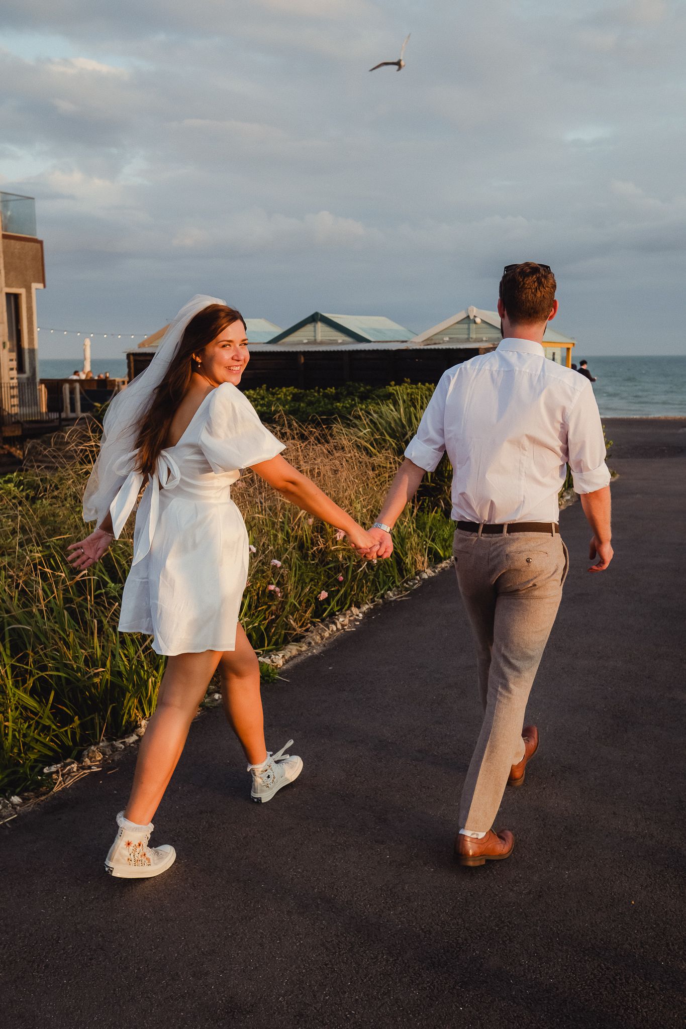 A couple in white outfits holding hands while walking along a coastal path at sunset.