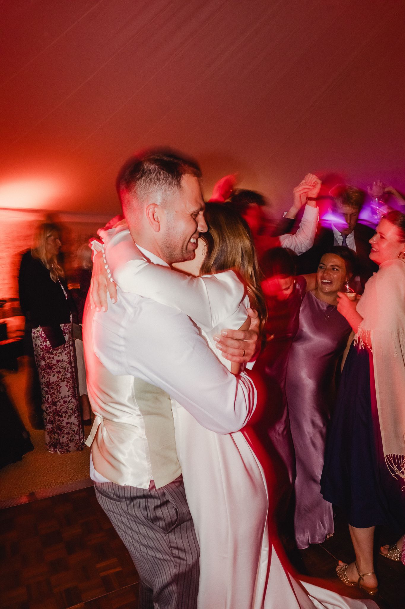 Wedding guests dance together under dramatic red lighting at an evening reception celebration.