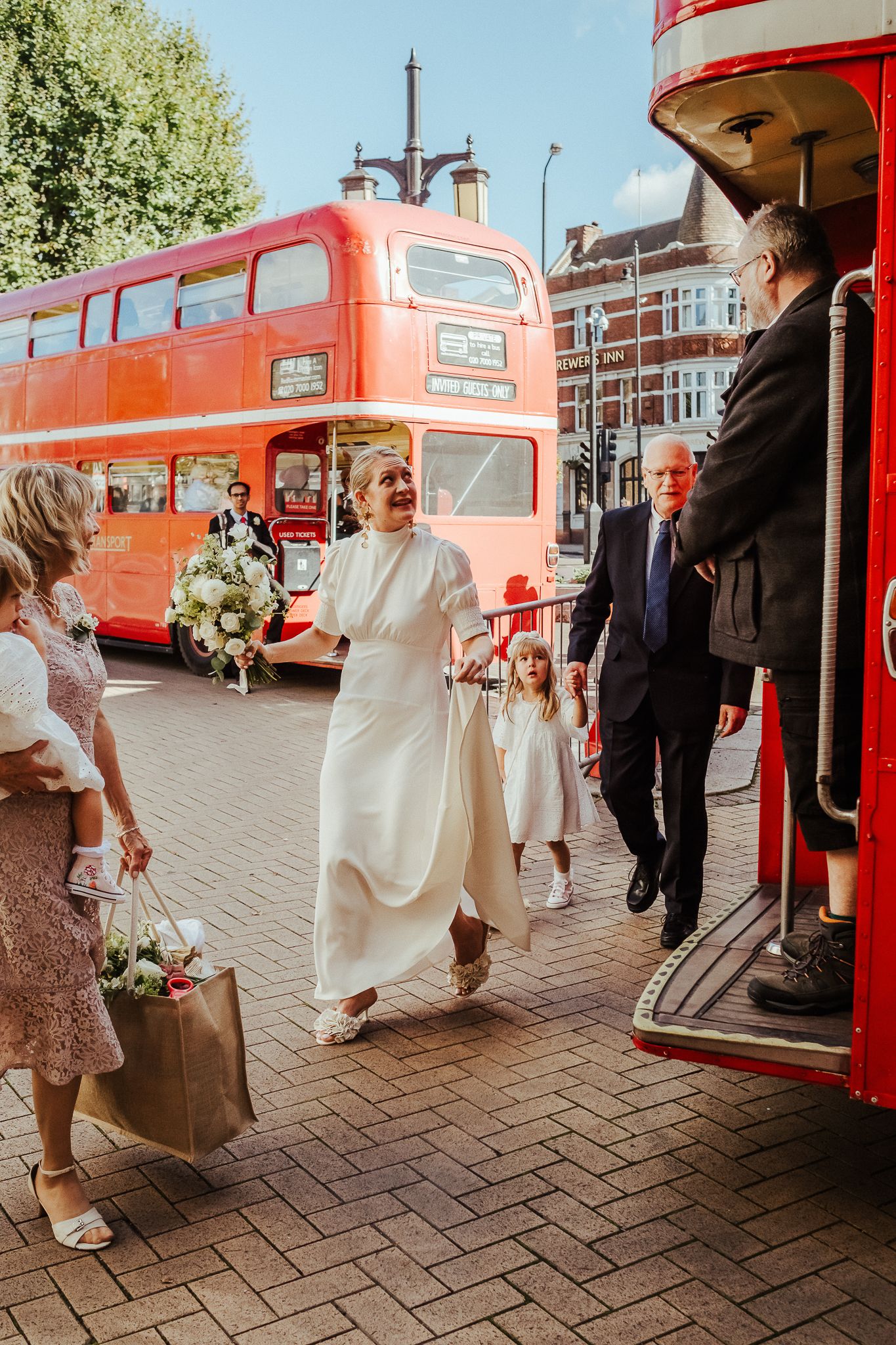 A vintage red double-decker bus serves as wedding transportation on a London street with wedding party boarding.