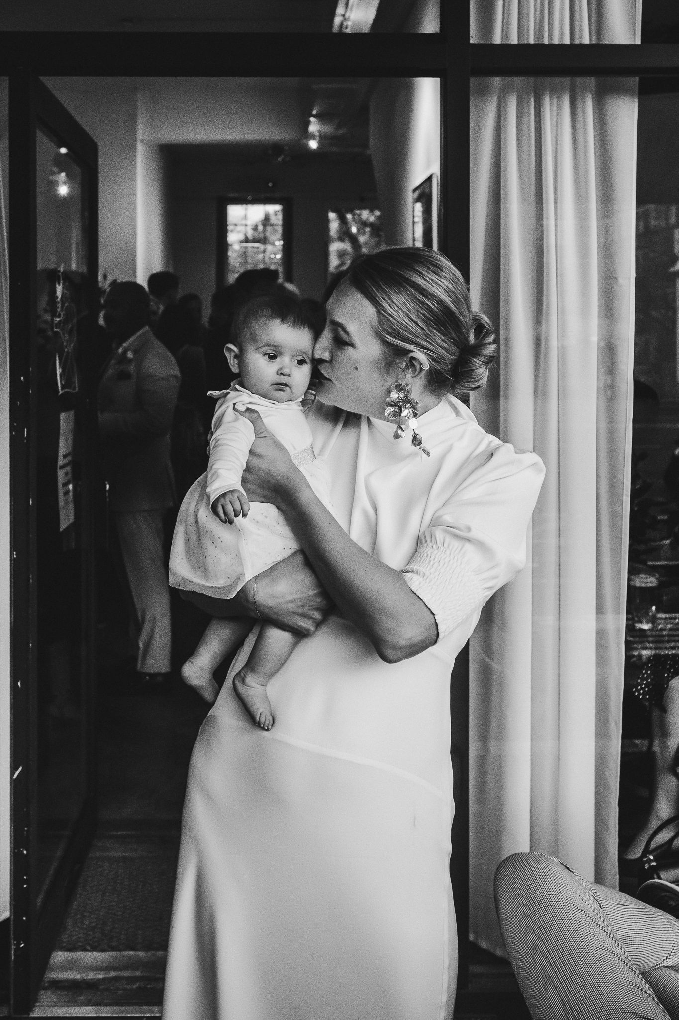 Black and white photo shows parent lifting toddler while standing among sheer curtains in softly lit room.