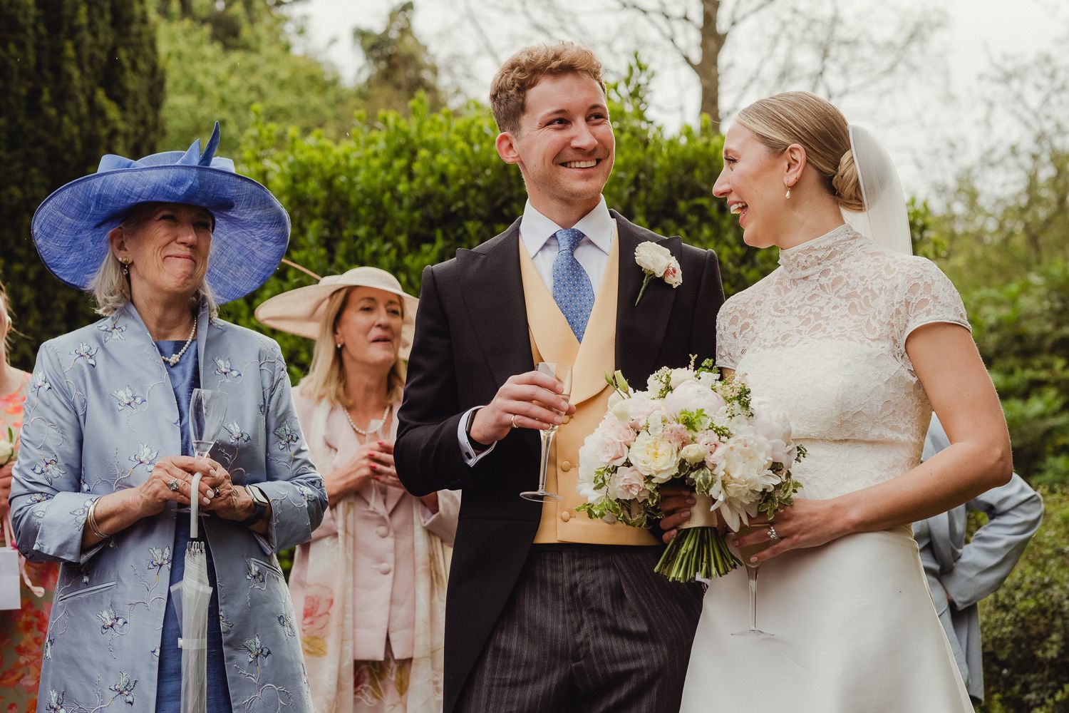 A joyful outdoor wedding scene with guests in formal attire including a blue hat, and a bouquet of white flowers.
