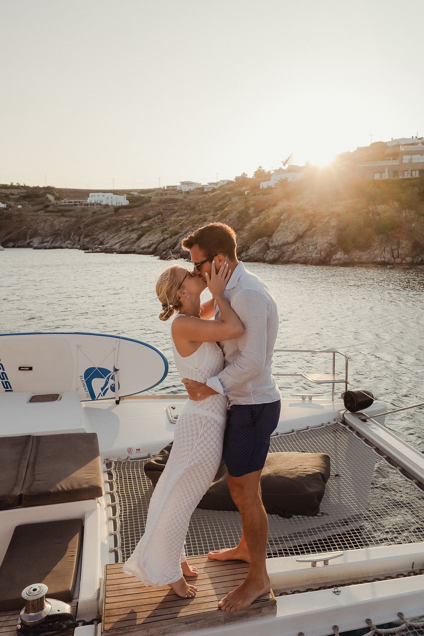 Romantic couple embraces on a catamaran deck at sunset near a rocky coastline.