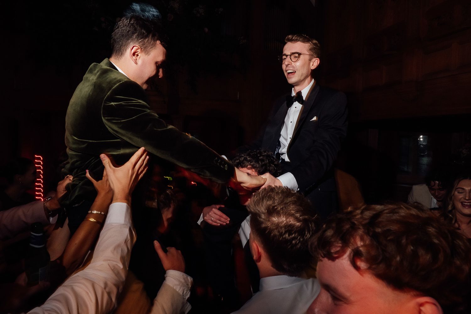 Two men in formal attire shaking hands energetically at a crowded wedding celebration.