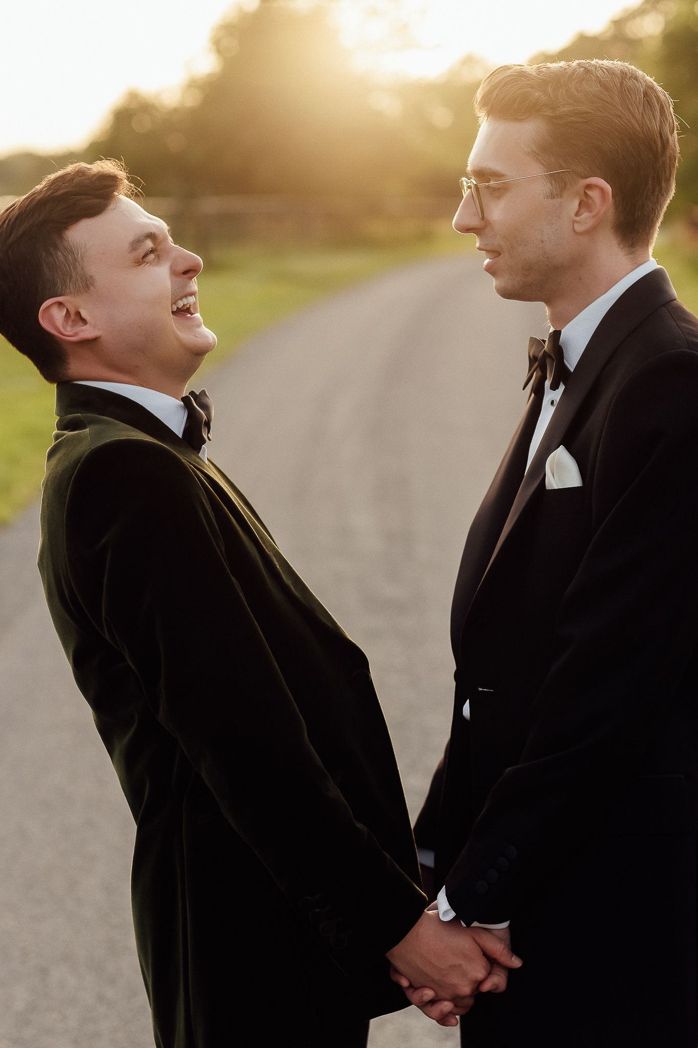 Two grooms in black tuxedos hold hands and share a joyful moment on a sunlit road at golden hour.